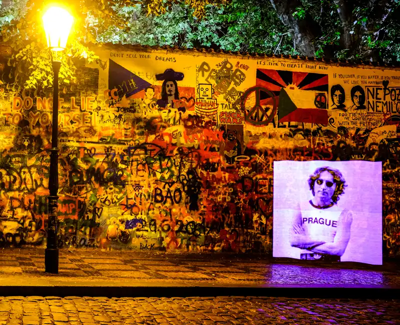 Night photo of the Lennon Wall with a long exposure shot, featuring a reimagined image of John Lennon wearing a white undershirt with the word 'Prague' printed on it. The iconic photo is recreated by Czech street artist moustache.