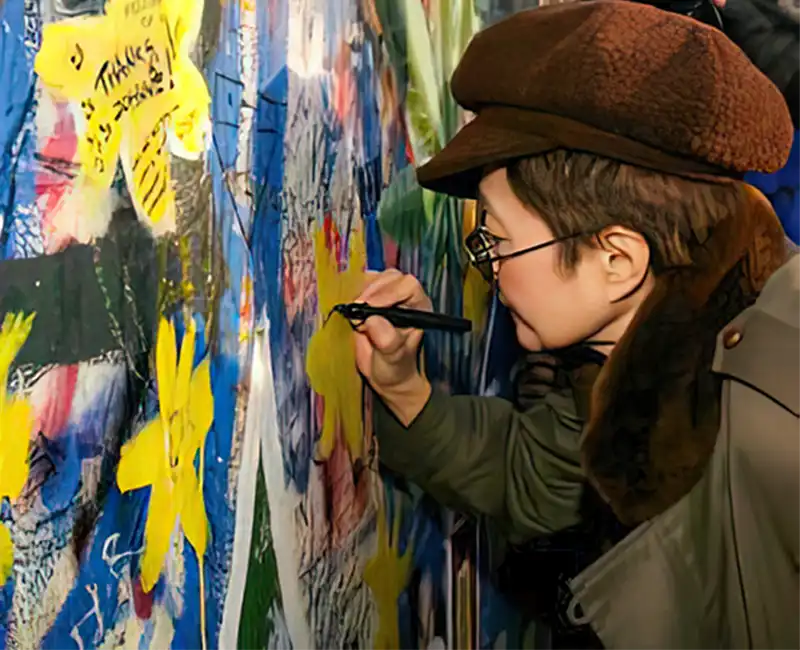 Yoko Ono signing her name on the Lennon Wall in Prague during a 2003 media event organized by the Kampa Museum. She also lit a candle and wrote heartfelt messages in small print, adding her personal touch to the wall's evolving history.